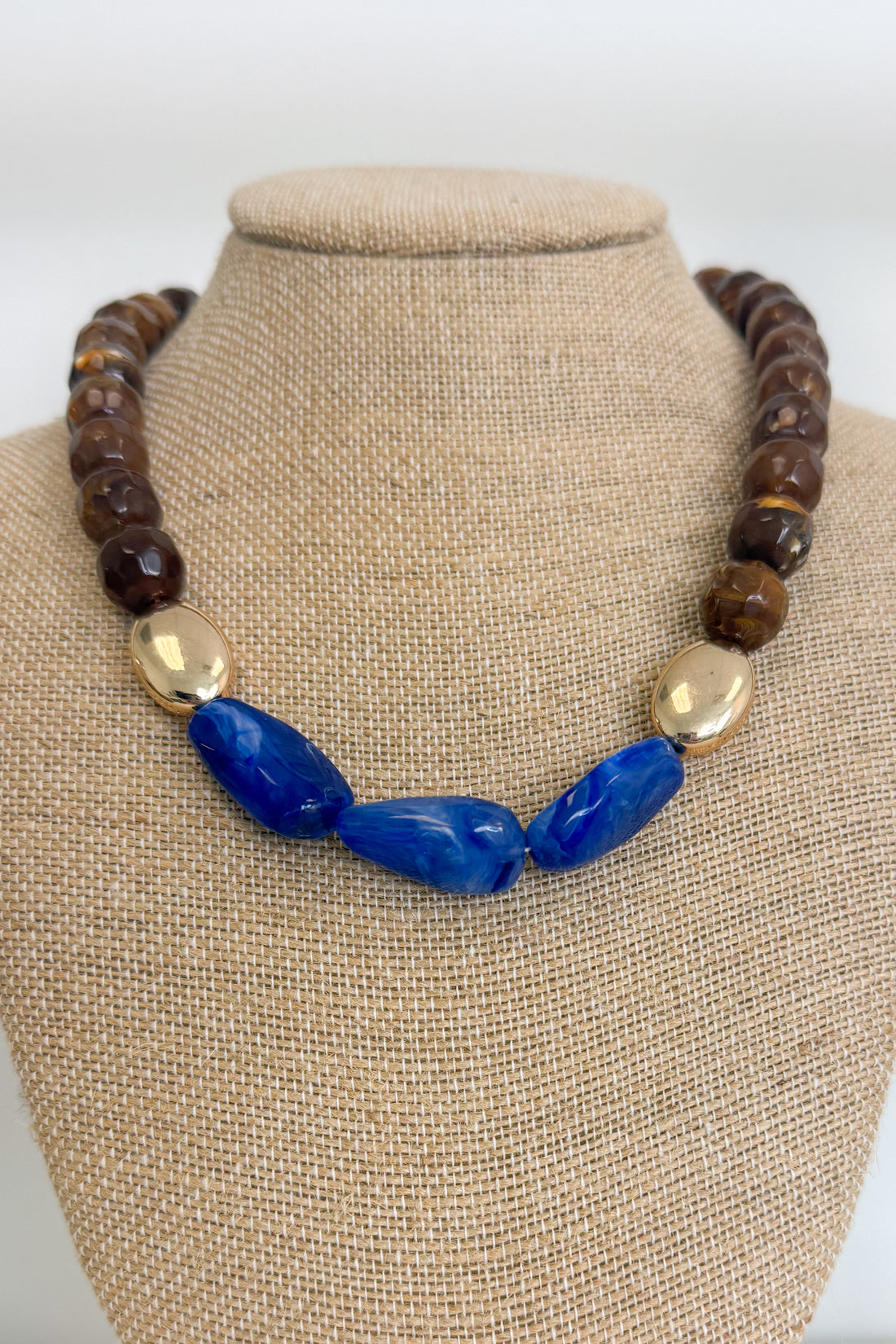 A statement necklace displayed on a beige fabric jewelry bust. The necklace features a strand of dark brown faceted beads, a central section of polished blue stone beads, and two metallic gold oval beads.