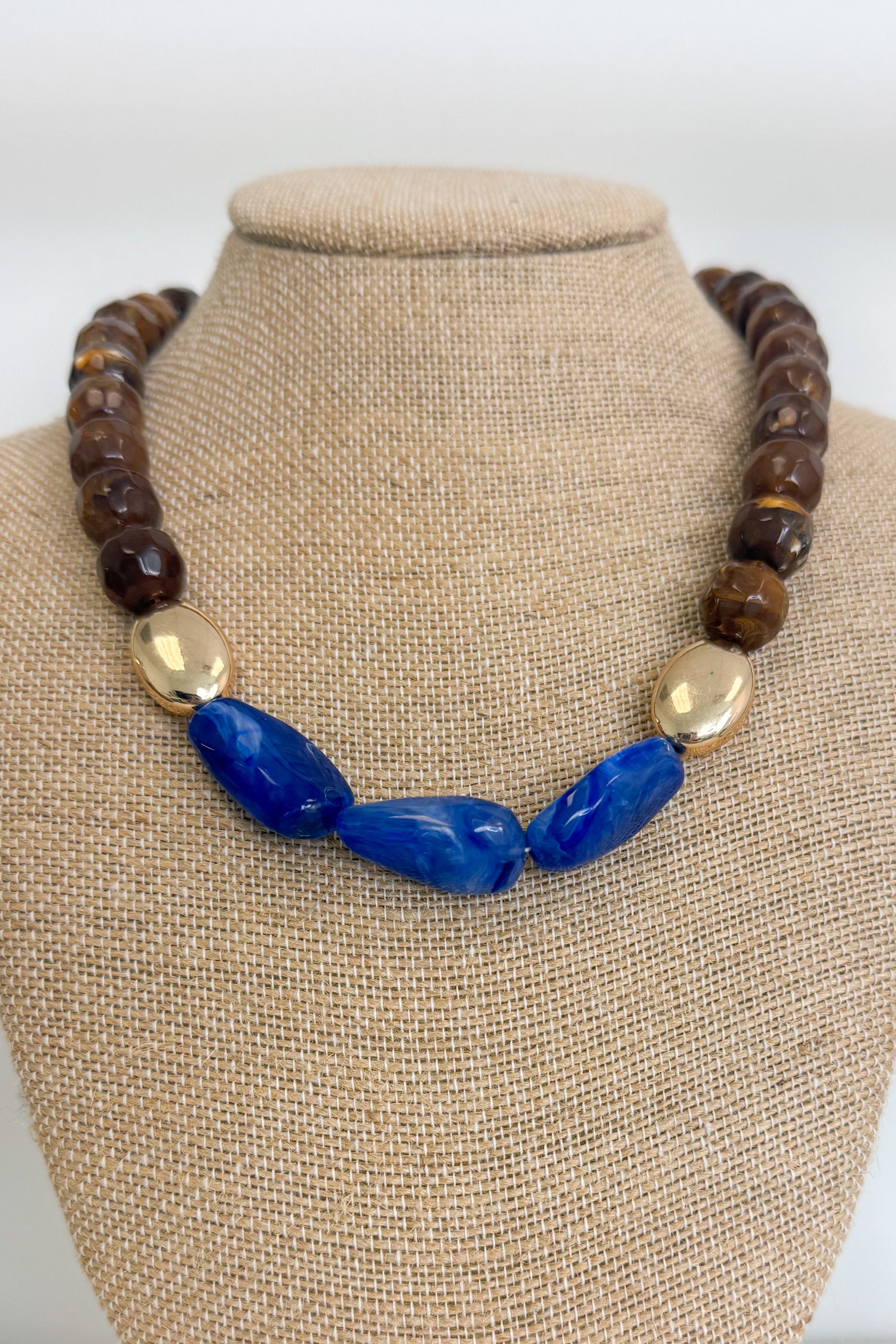 A statement necklace displayed on a beige fabric jewelry bust. The necklace features a strand of dark brown faceted beads, a central section of polished blue stone beads, and two metallic gold oval beads.