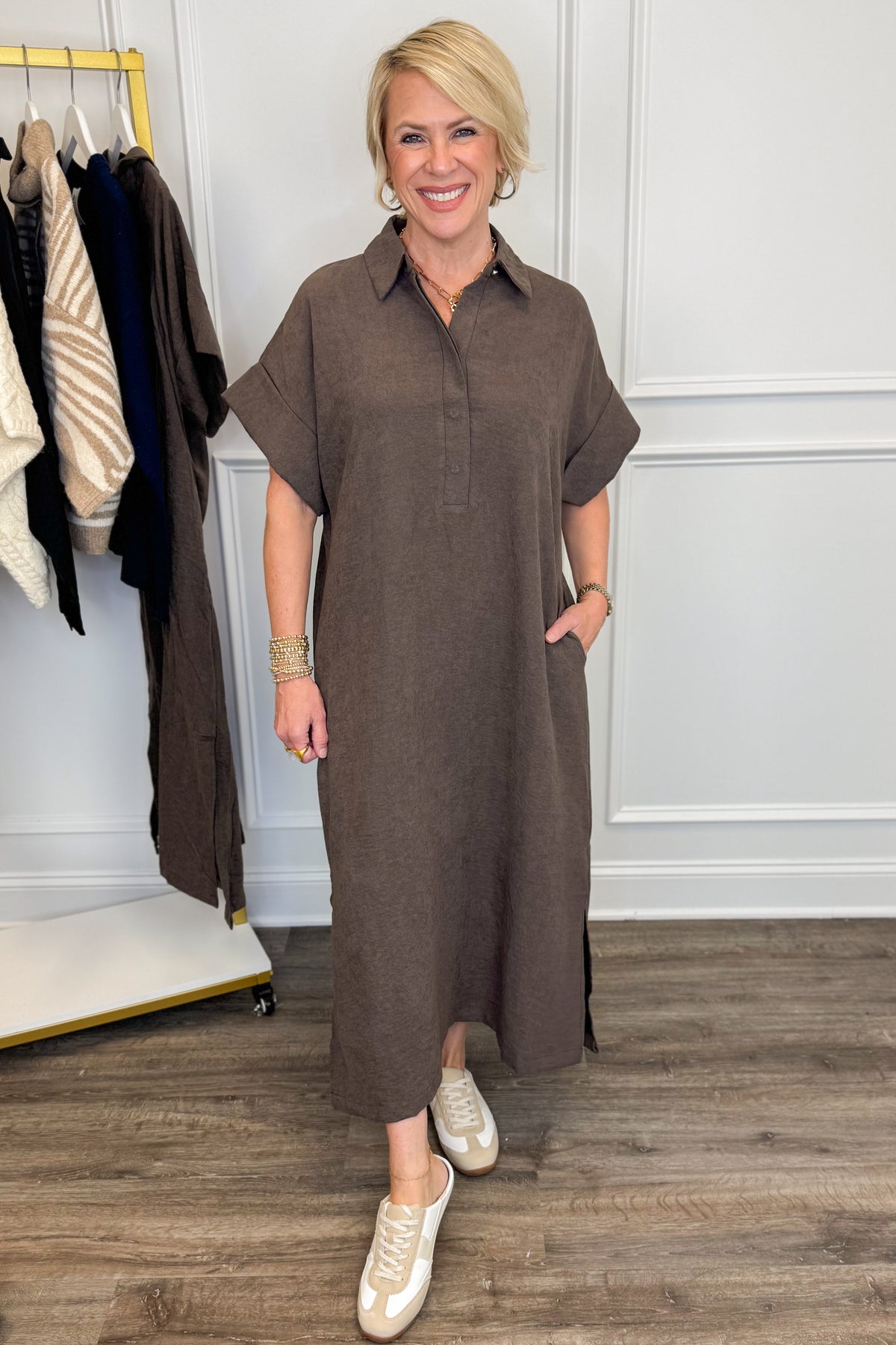 A woman modeling a brown short-sleeve collared midi dress with a button placket and side pockets, standing in a boutique setting with a clothing rack and wood floor visible.