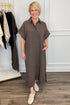 A woman modeling a brown short-sleeve collared midi dress with a button placket and side pockets, standing in a boutique setting with a clothing rack and wood floor visible.