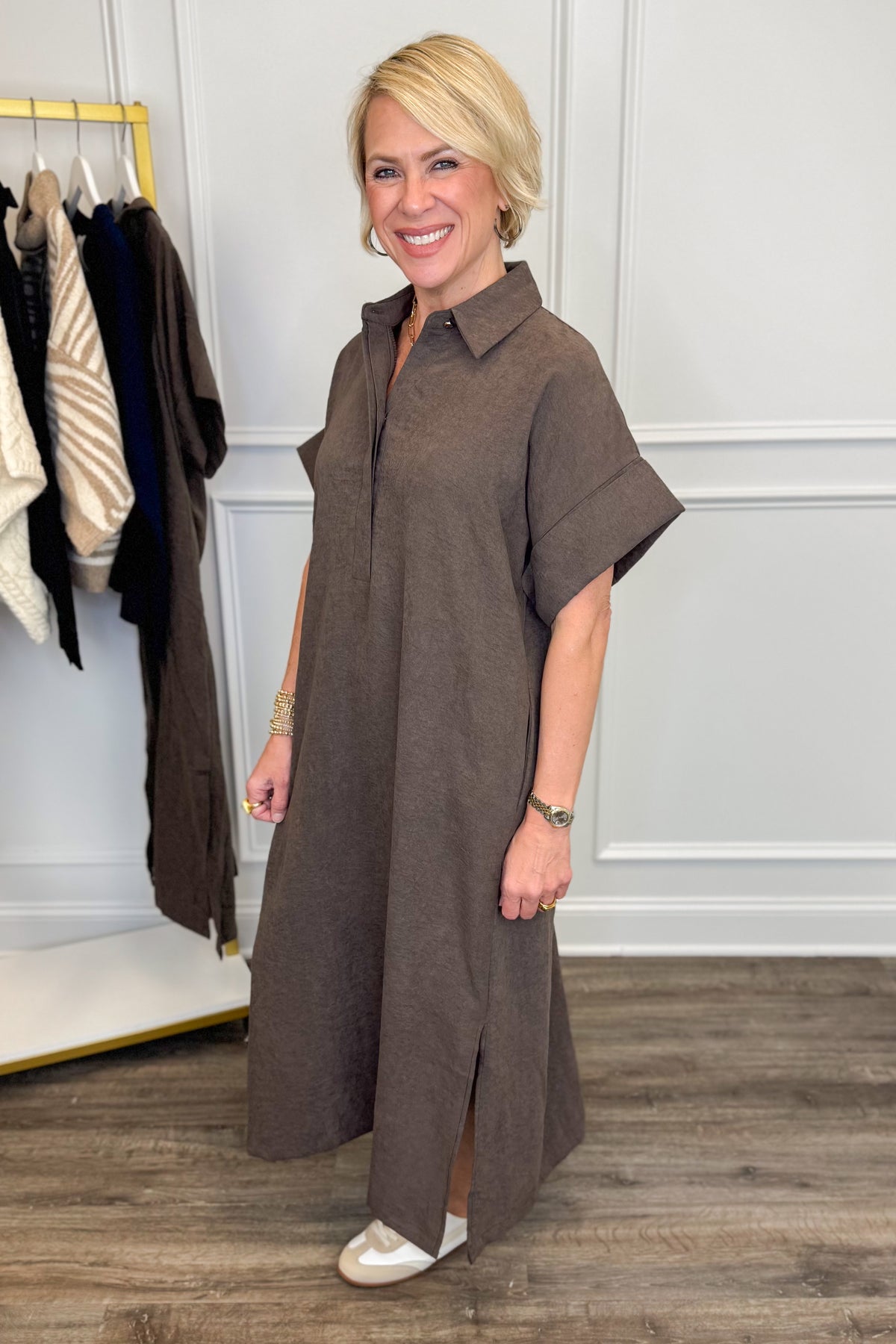 A woman modeling a brown short-sleeve collared dress in a boutique setting with a clothing rack and wood floor visible.