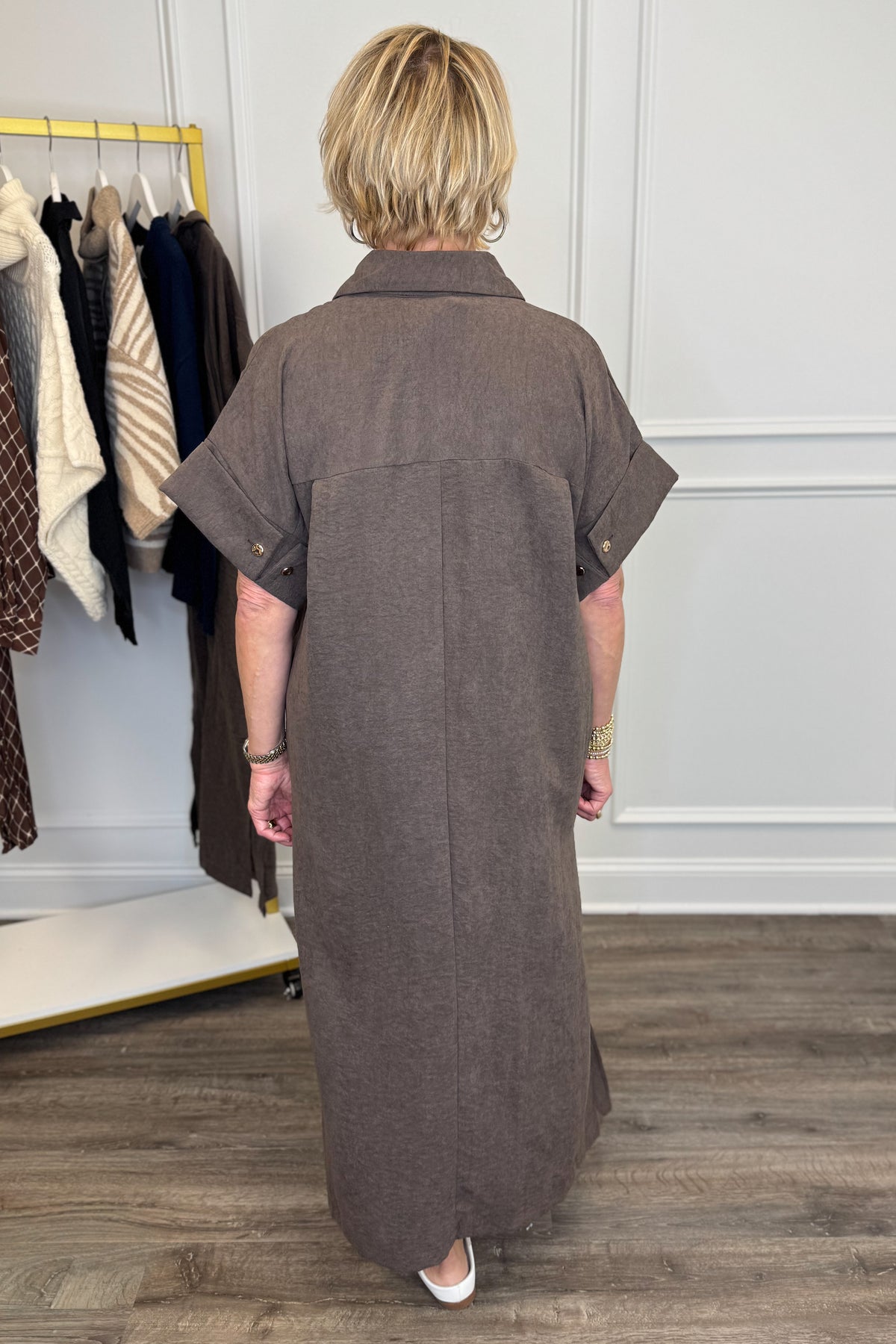A woman viewed from behind wearing a brown short-sleeve collared shirt dress in a boutique setting with a clothing rack and wood floor.
