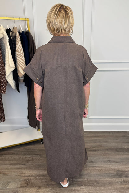 A woman viewed from behind wearing a brown short-sleeve collared shirt dress in a boutique setting with a clothing rack and wood floor.