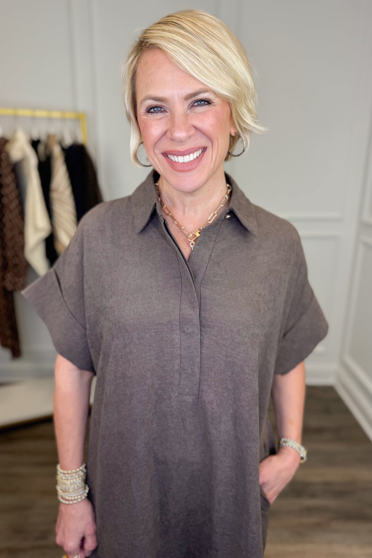 A woman modeling a brown short-sleeve collared midi dress with a button placket and side pockets, standing in a boutique setting.