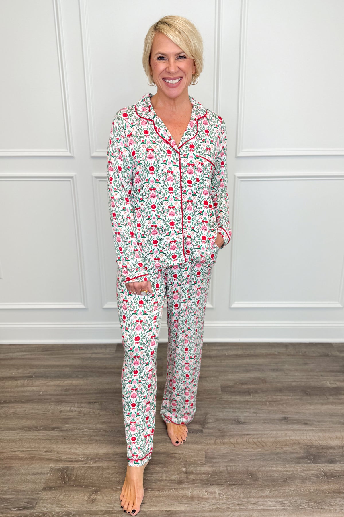 Model wearing the Trimmed With Joy Charlotte Pajama Set, featuring a festive red, pink, and green holiday floral print on a white background. The set includes a long-sleeve button-down top with red contrast piping, a chest pocket, and matching straight-leg pajama pants.