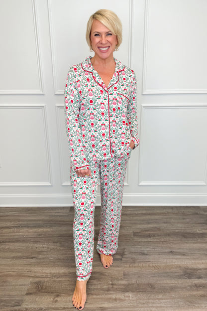 Model wearing the Trimmed With Joy Charlotte Pajama Set, featuring a festive red, pink, and green holiday floral print on a white background. The set includes a long-sleeve button-down top with red contrast piping, a chest pocket, and matching straight-leg pajama pants.