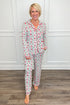 Model wearing the Trimmed With Joy Charlotte Pajama Set, featuring a festive red, pink, and green holiday floral print on a white background. The set includes a long-sleeve button-down top with red contrast piping, a chest pocket, and matching straight-leg pajama pants.