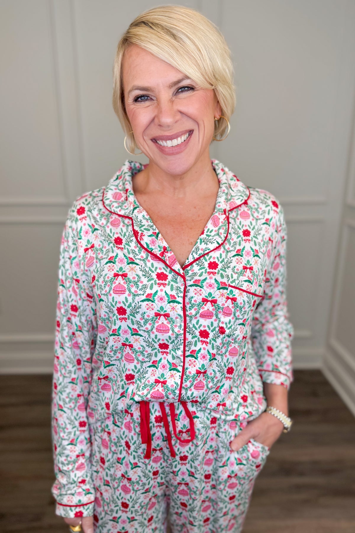 Model wearing the Trimmed With Joy Charlotte Pajama Set, featuring a festive red, pink, and green holiday floral print on a white background. The set includes a long-sleeve button-down top with red contrast piping, a chest pocket, and matching straight-leg pajama pants.