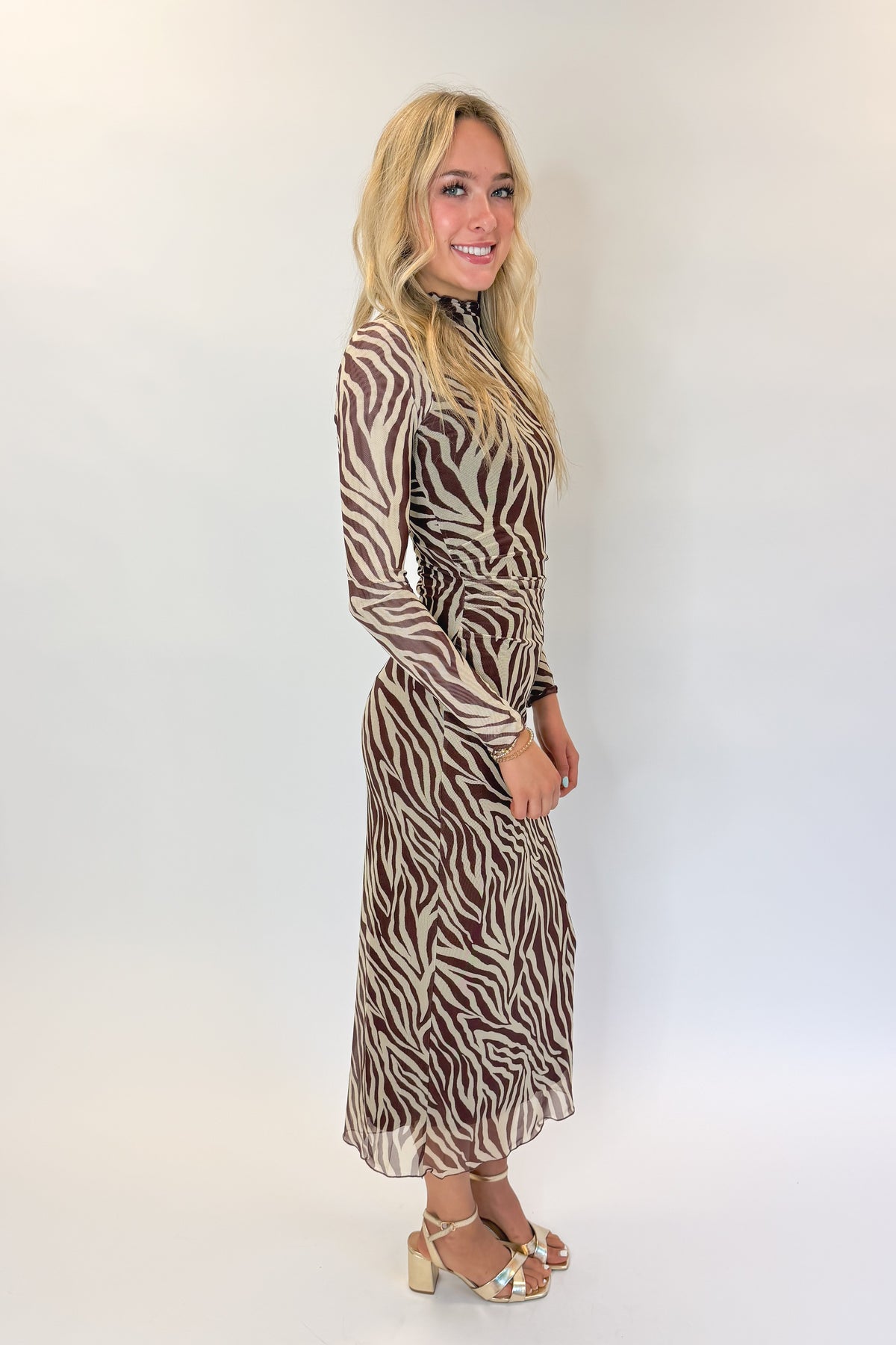 Side view of woman wearing Swagger Boutique’s Velvet Rouge Sevilla Zebra Mesh Midi Dress by Z Supply with heels against white studio background.