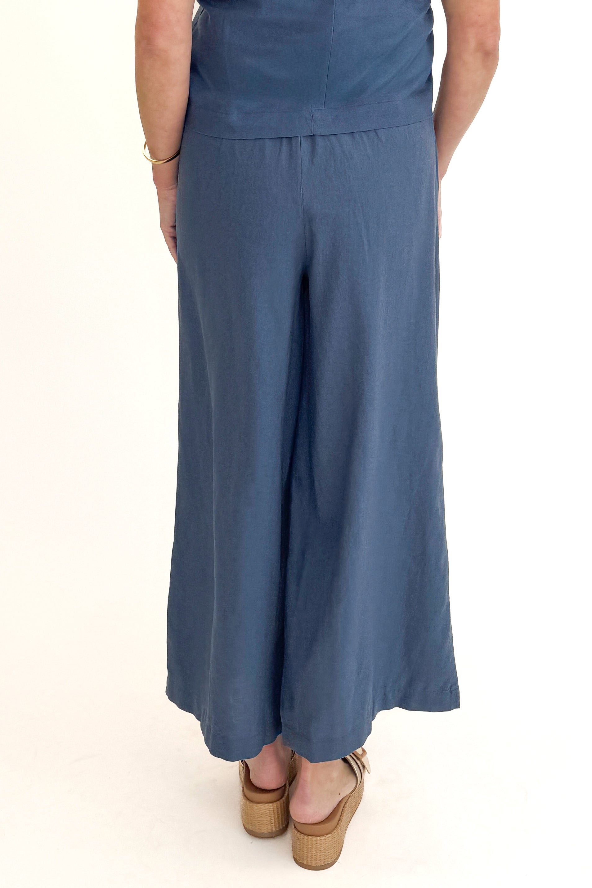 Back view of woman wearing Vintage Indigo Scout Linen Pants by Z Supply with matching Vintage Indigo Sloane Linen Top by Z Supply against white studio background.
