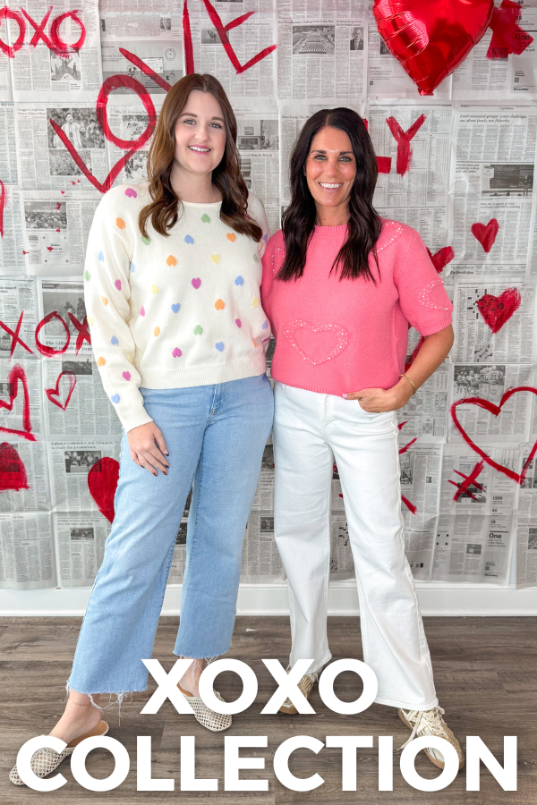 Two women standing in front of a decorated wall with 'XOXO' and heart shapes, wearing colorful clothing.