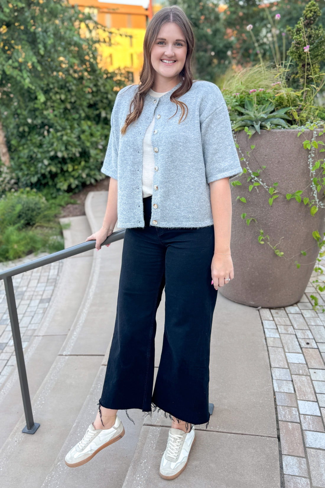 Woman standing outdoors wearing a light blue jacket, white shirt, dark blue pants, and white sneakers.