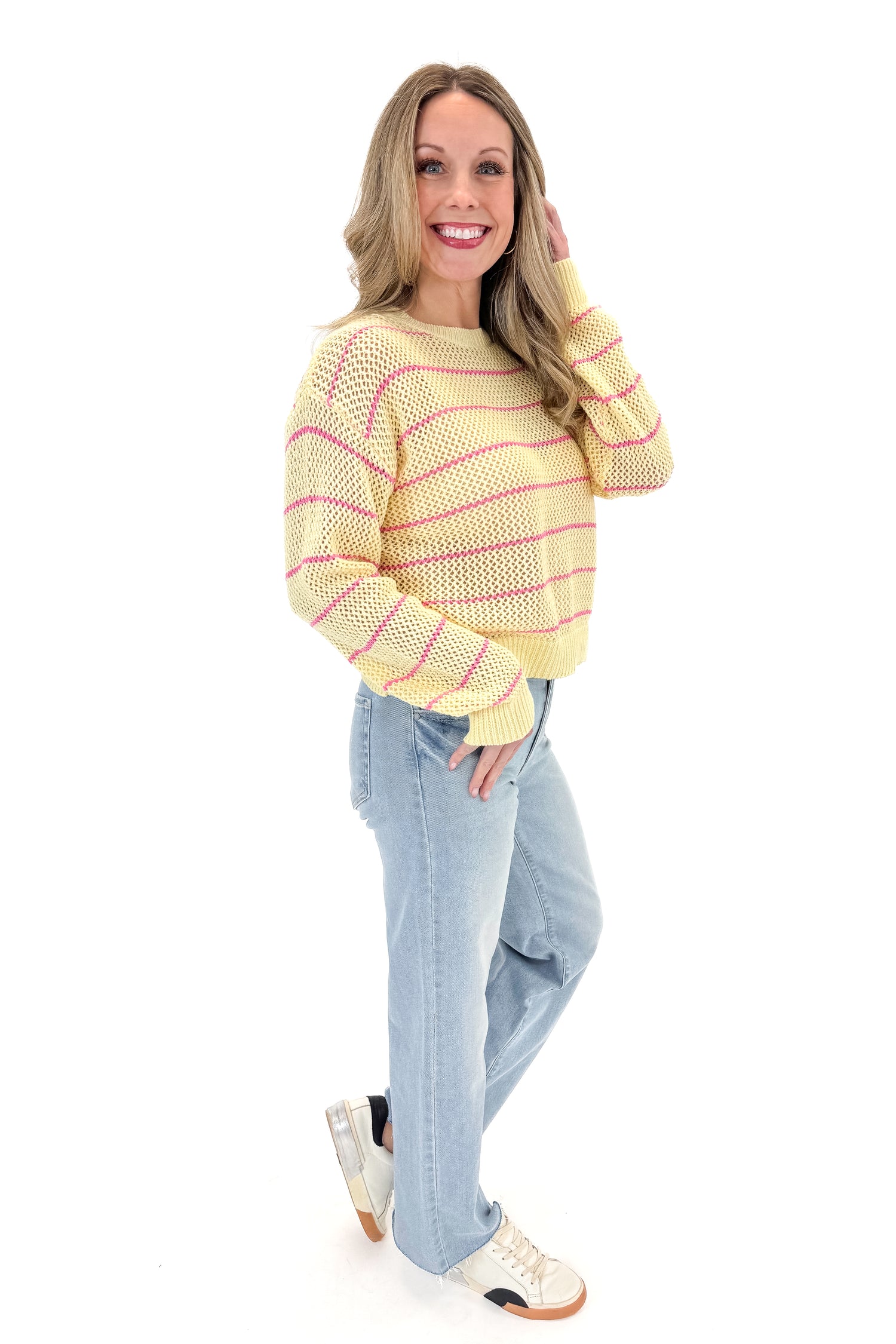 Side view of woman wearing Yellow &amp; Pink Striped Loose Knit Sweater with jeans against white studio background.