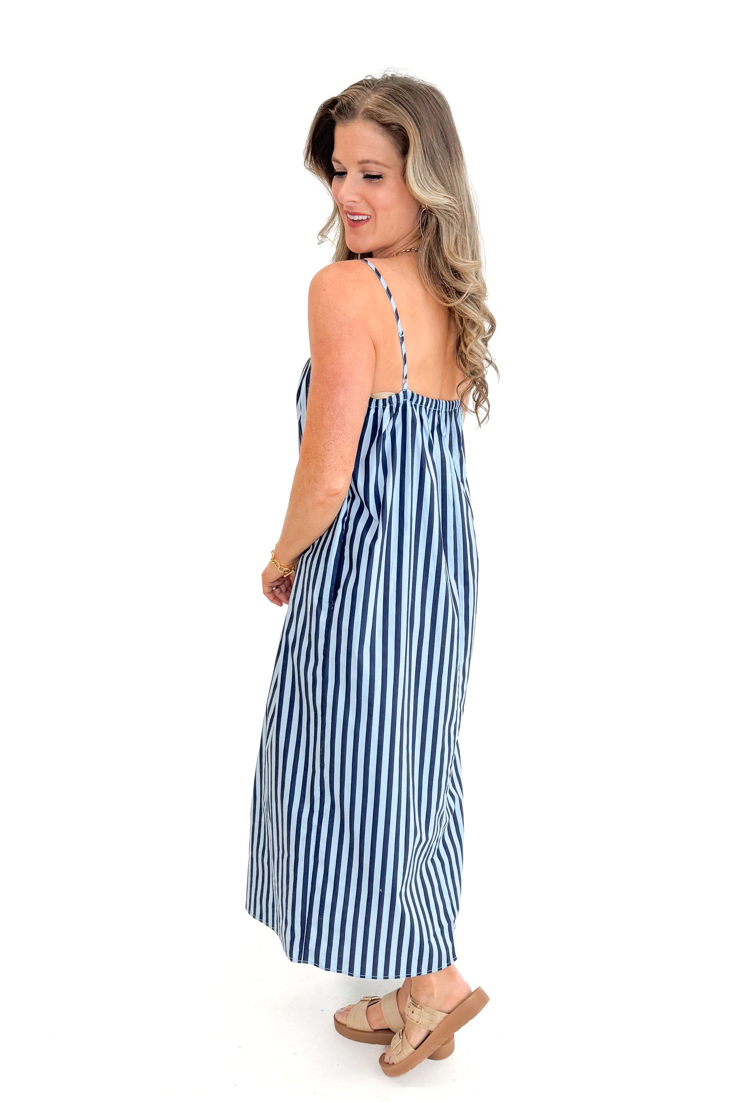 Side view of woman wearing Blue &amp; Navy Striped Midi Dress with sandals against white studio background.