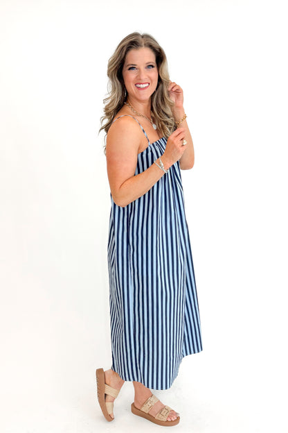Side view of woman wearing Blue &amp; Navy Striped Midi Dress with sandals against white studio background.