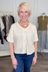 A smiling blonde woman stands in a boutique wearing a short-sleeve cream knit top with buttons, blue jeans, and layered gold-toned jewelry. Clothing racks and a mannequin are visible in the softly lit store interior.