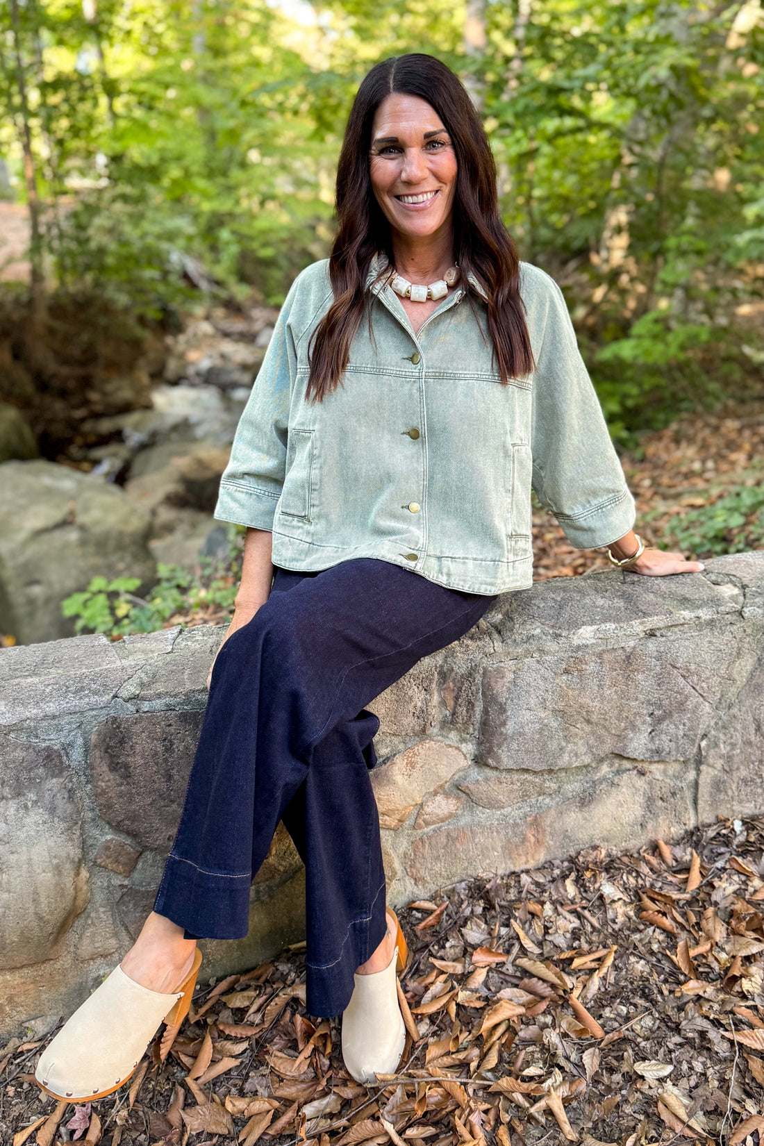 Front view of woman wearing Swagger Boutique’s Washed Olive Utility Jacket with jeans sitting outside at a park.