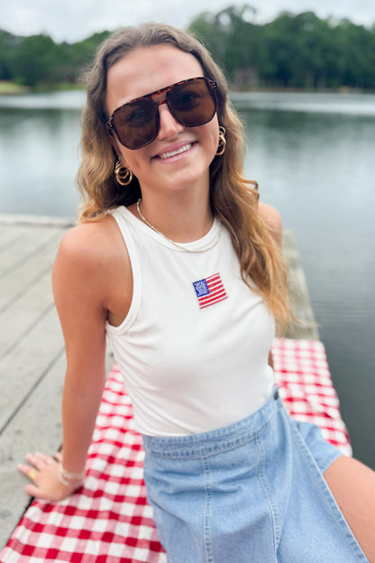 Front view of woman wearing Swagger Boutique’s White Beaded Flag Tank with jean skort sitting outside at the lake.