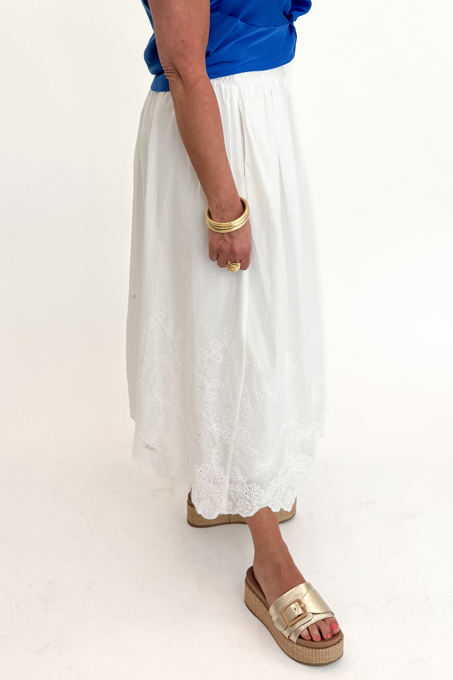 Side view of woman wearing Swagger Boutique’s Blue Sleeveless Tank With Twist by Skies Are Blue with White Embroidered Skirt against white studio background.