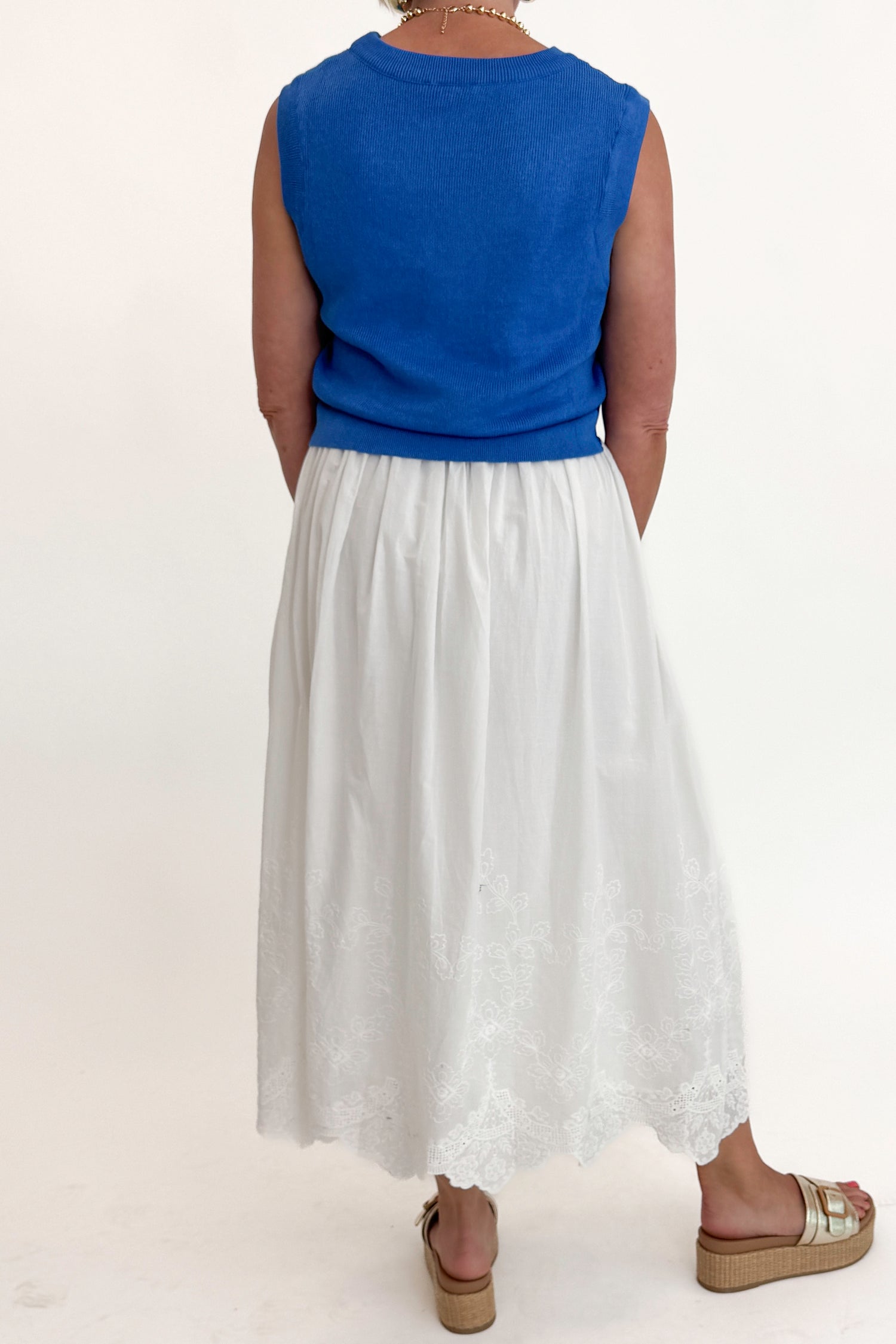 Back view of woman wearing Swagger Boutique’s Blue Sleeveless Tank With Twist by Skies Are Blue with White Embroidered Skirt against white studio background.