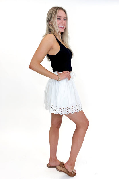 Side view of woman wearing Swagger Boutique’s White Eyelet Skort with black tank against white studio background.