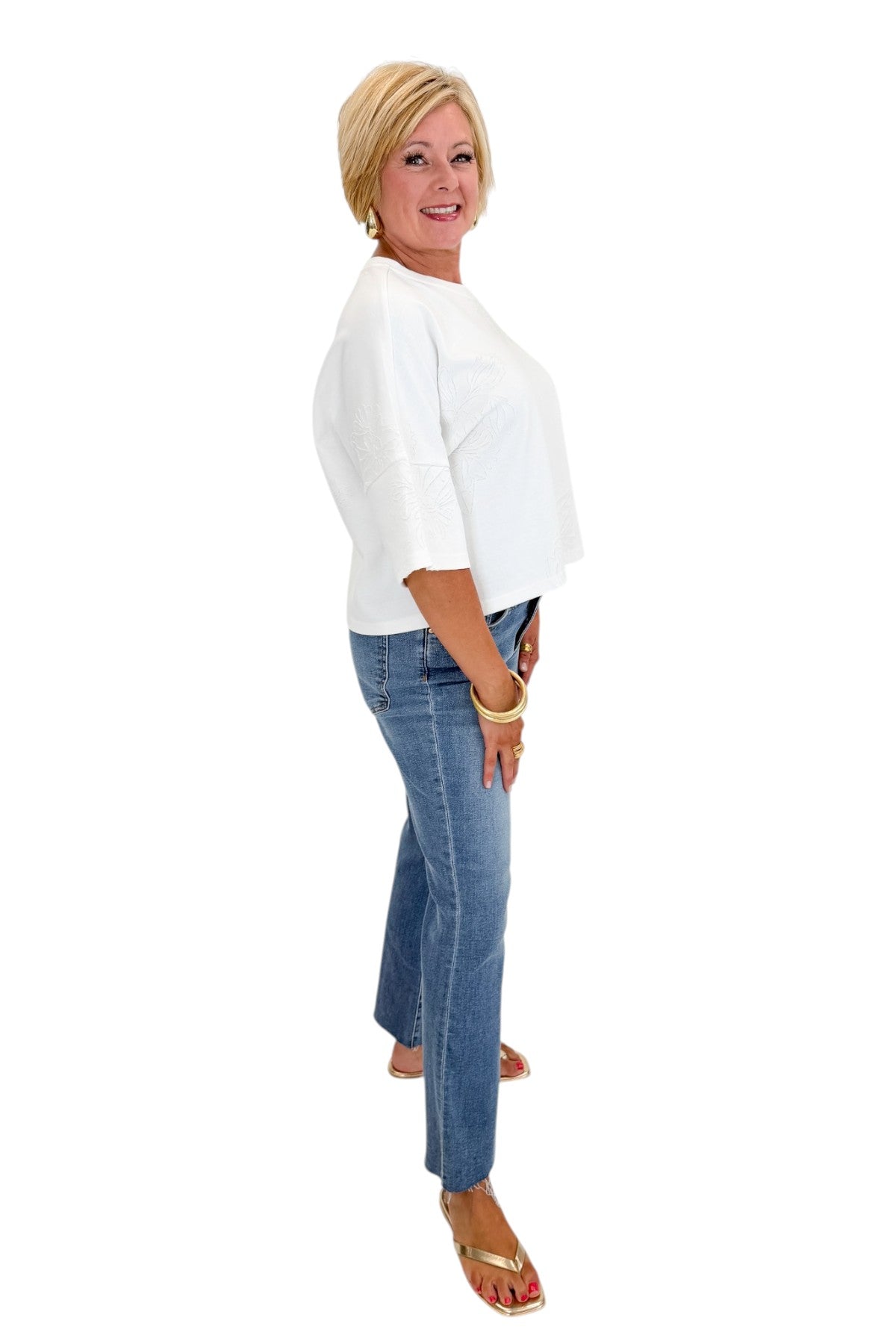 Side view of woman wearing Swagger Boutique’s White Floral Embroidered Short Sleeve Sweatshirt by Entro with jeans against white studio background.