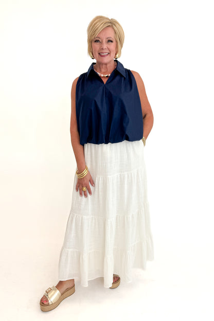 Front view of woman wearing Swagger Boutique’s White Flowy Tiered Cotton Skirt with blue top against white studio background.