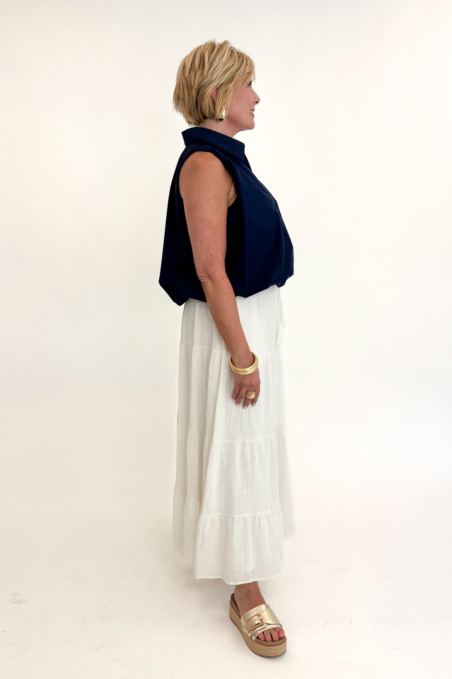 Side view of woman wearing Swagger Boutique’s White Flowy Tiered Cotton Skirt with blue top against white studio background.