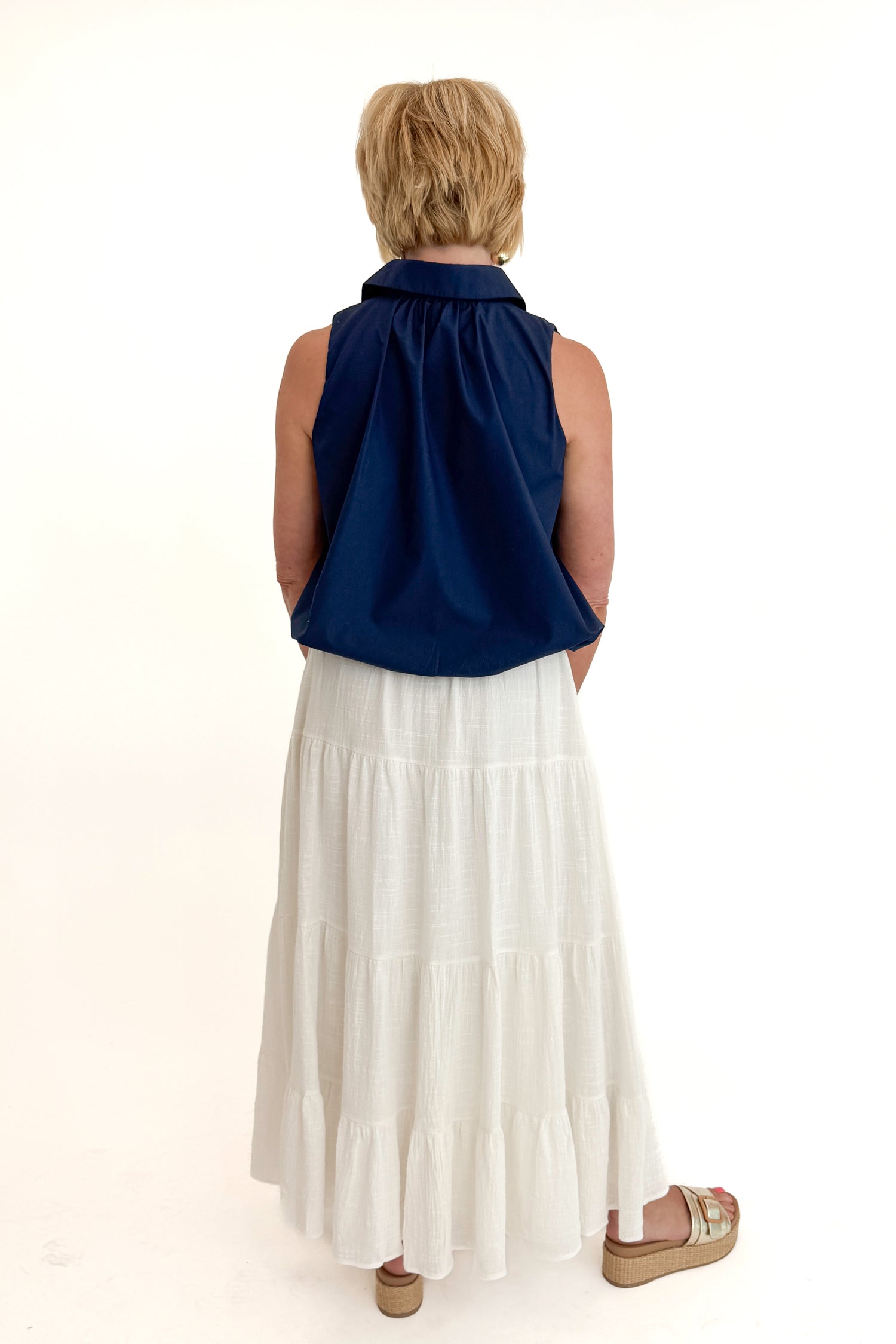 Back view of woman wearing Swagger Boutique’s White Flowy Tiered Cotton Skirt with blue top against white studio background.