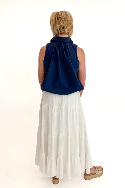 Back view of woman wearing Swagger Boutique’s White Flowy Tiered Cotton Skirt with blue top against white studio background.