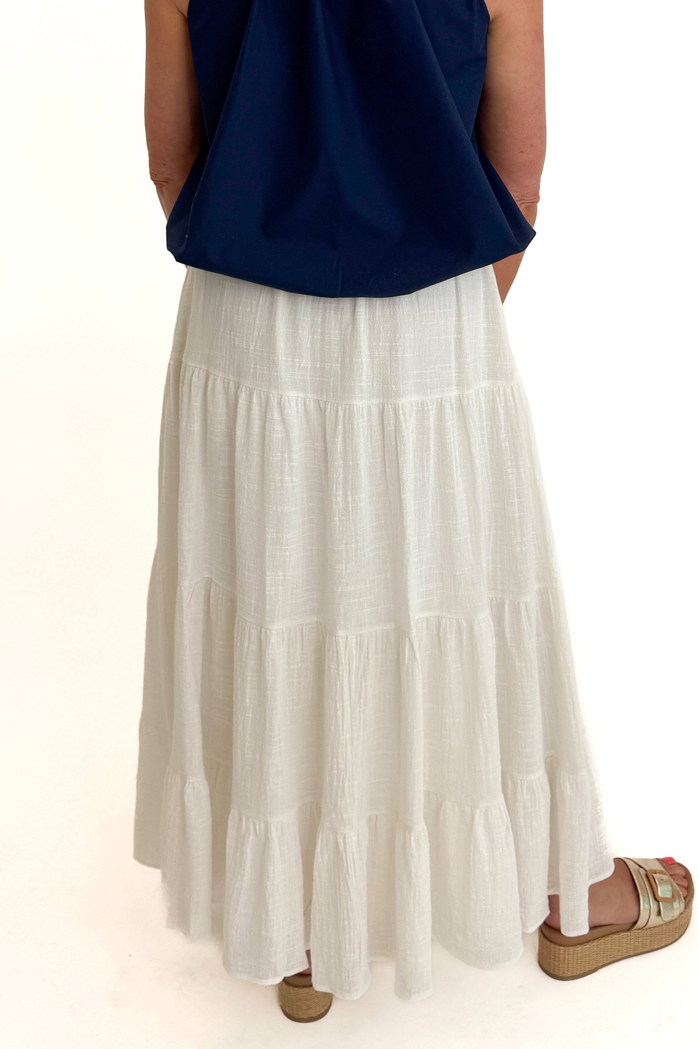 Back detail view of woman wearing Swagger Boutique’s White Flowy Tiered Cotton Skirt with blue top against white studio background.