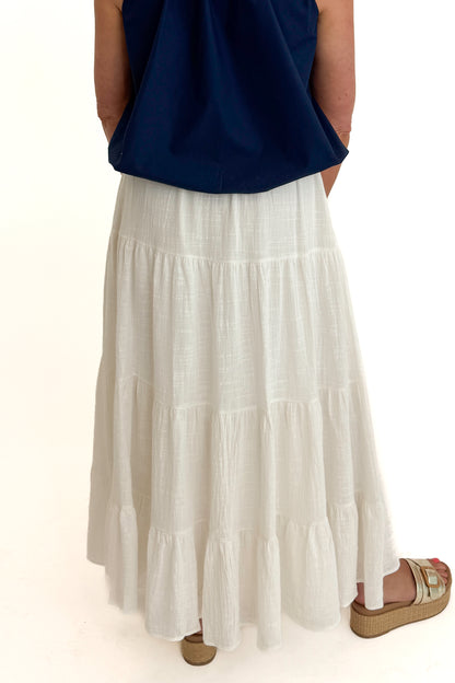 Back detail view of woman wearing Swagger Boutique’s White Flowy Tiered Cotton Skirt with blue top against white studio background.