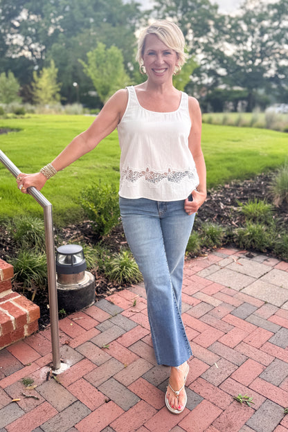 Front view of woman wearing Swagger Boutique’s White Lace Embroidered Detail Tank with jeans standing on a brick patio.