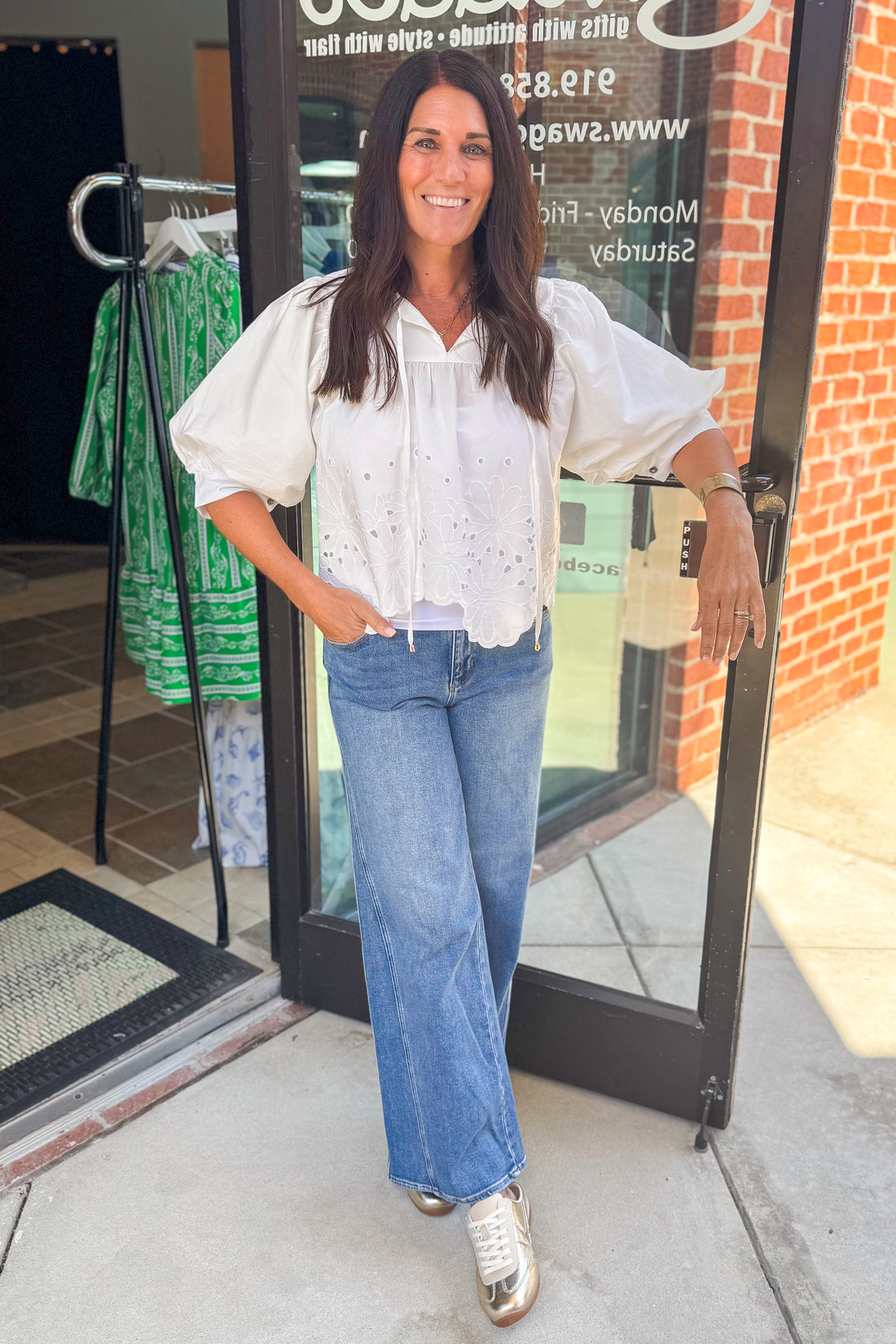 Front view of woman wearing Swagger Boutique’s White Puff Sleeve Eyelet Top with jeans standing outside the store.