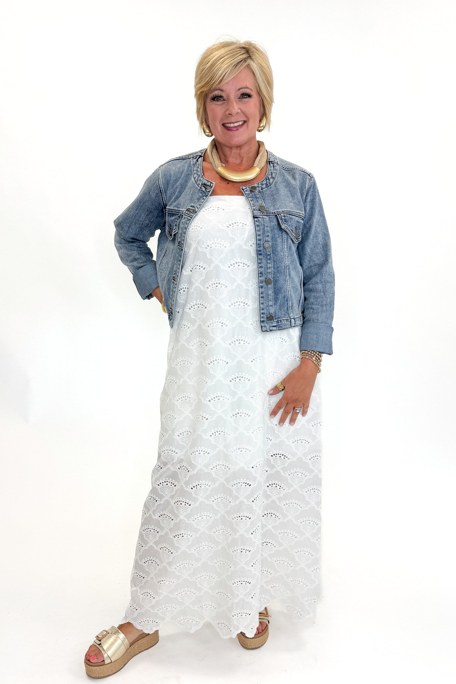 Front view of woman wearing Swagger Boutique’s White Strapless Eyelet Midi Dress by Entro and Medium Wash Collarless Denim Jacket by Just Black Denim with sandals against white studio background.