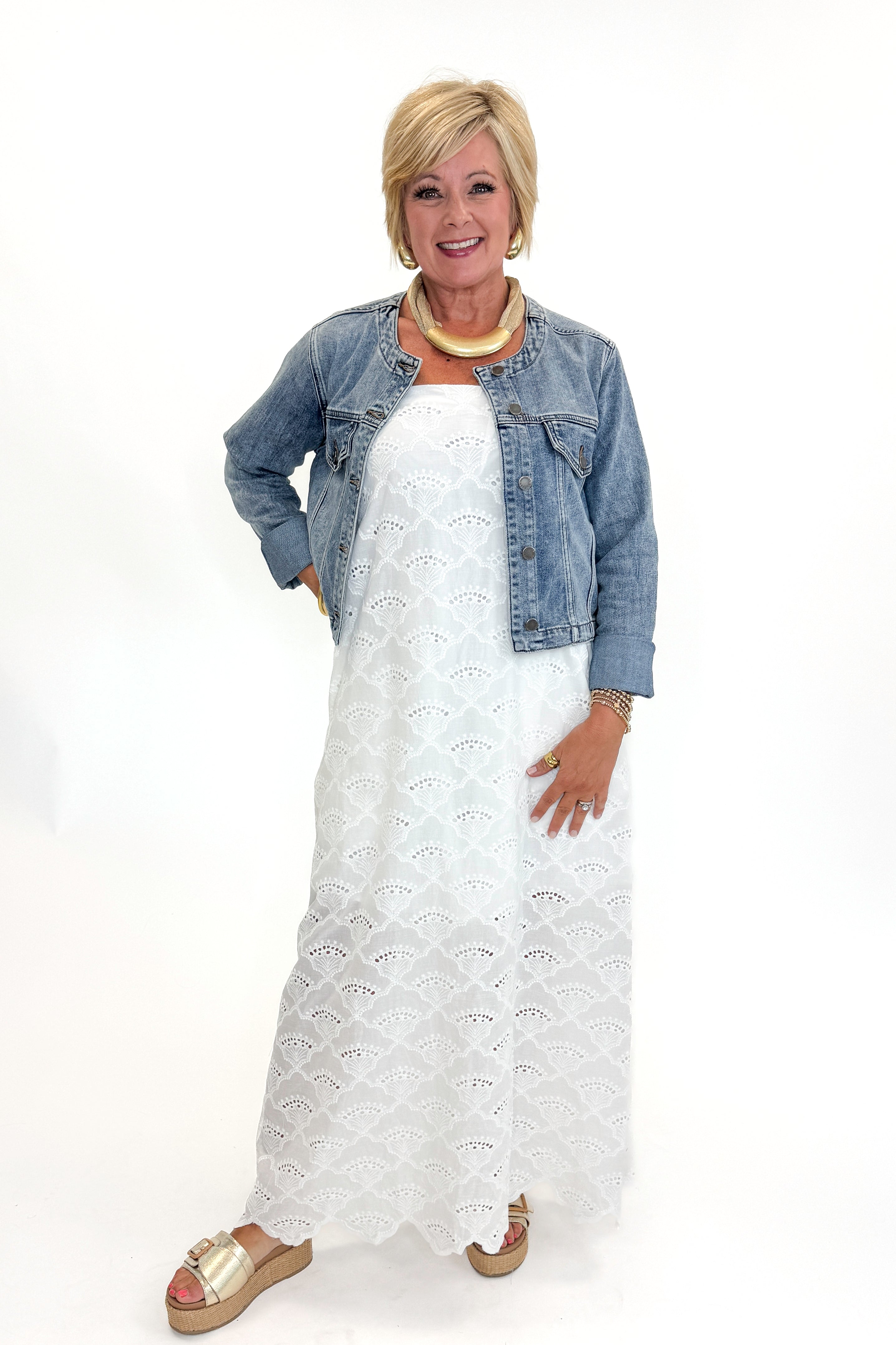 Front view of woman wearing Swagger Boutique’s White Strapless Eyelet Midi Dress by Entro and Medium Wash Collarless Denim Jacket by Just Black Denim with sandals against white studio background.