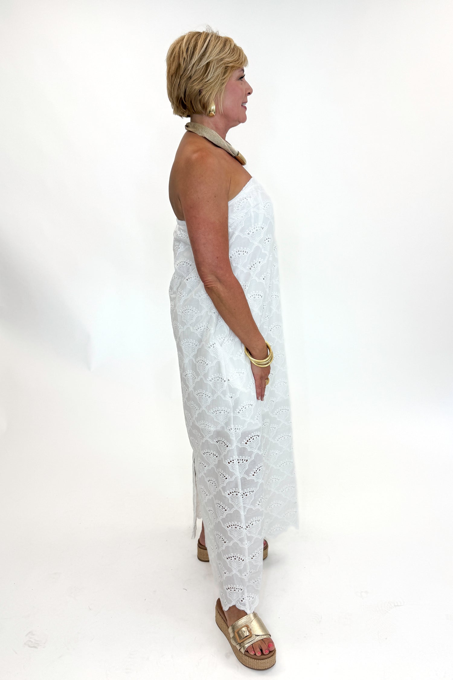 Side view of woman wearing Swagger Boutique’s White Strapless Eyelet Midi Dress by Entro with sandals against white studio background.