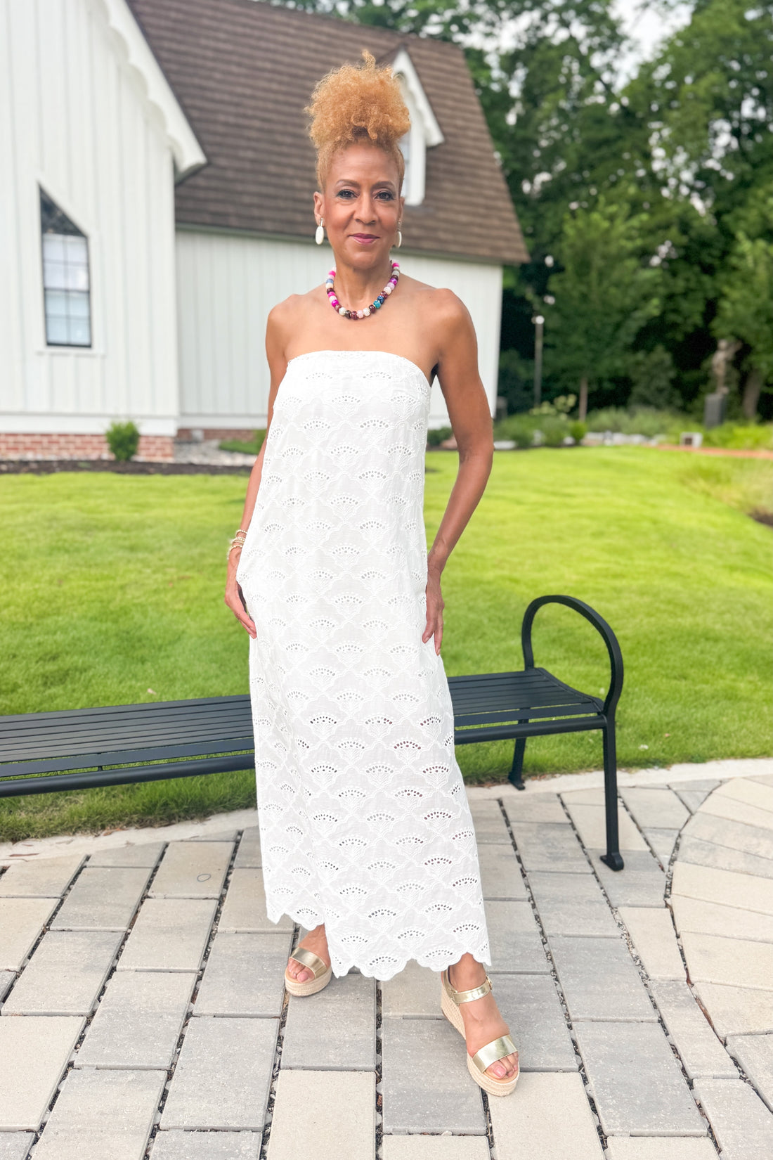 Front view of woman wearing Swagger Boutique’s White Strapless Eyelet Midi Dress by Entro with sandals standing outside by a bench.