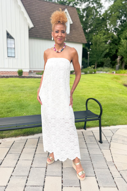 Front view of woman wearing Swagger Boutique’s White Strapless Eyelet Midi Dress by Entro with sandals standing outside by a bench.