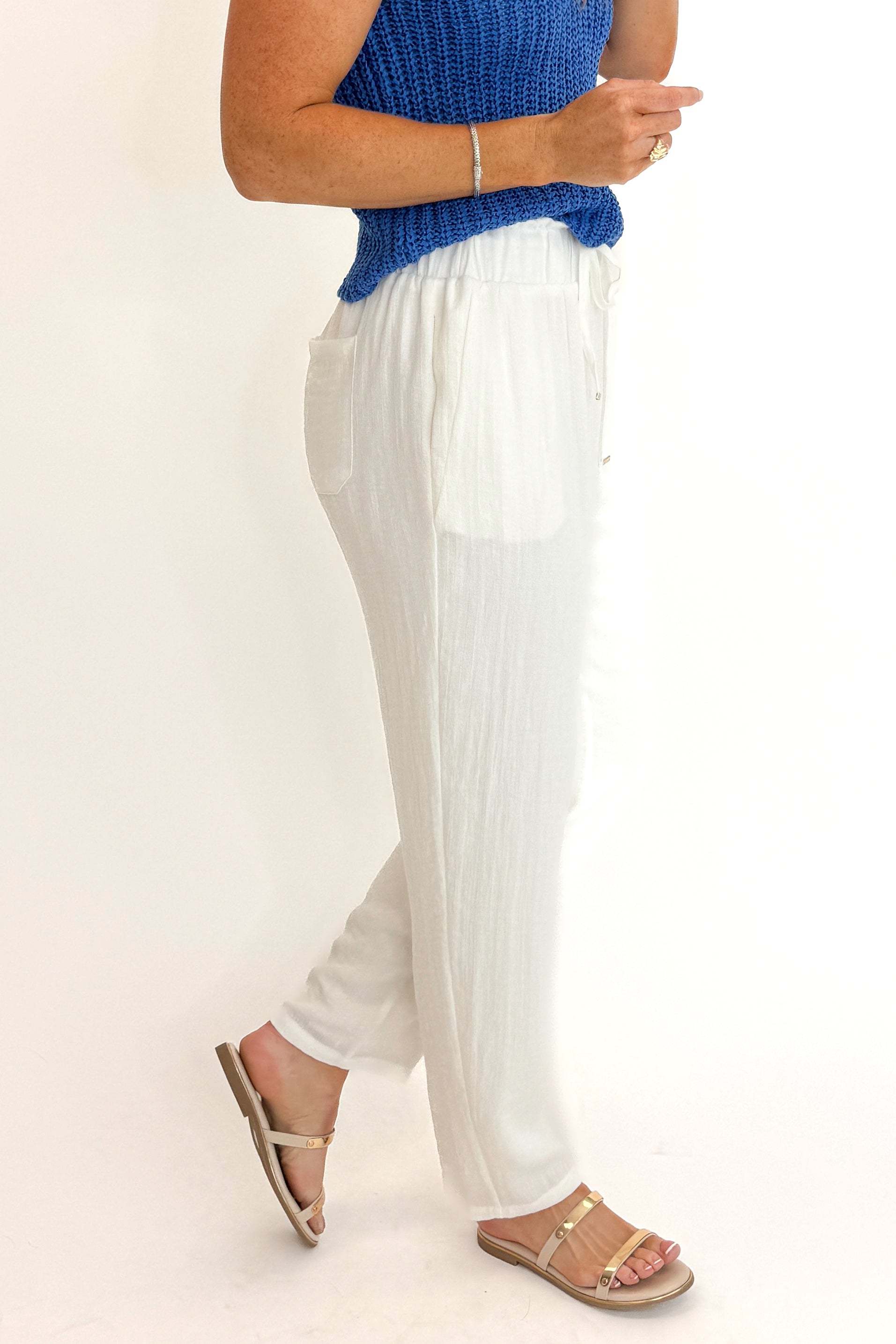 Side view of woman wearing Swagger Boutique’s White Woven Pants with blue tank against white studio background.