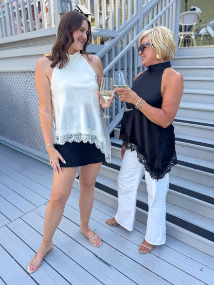 Front view of woman wearing Swagger Boutique’s Black Asymmetrical Halter Neck Satin Lace Top with jeans standing with woman wearing the white version and skirt outside.