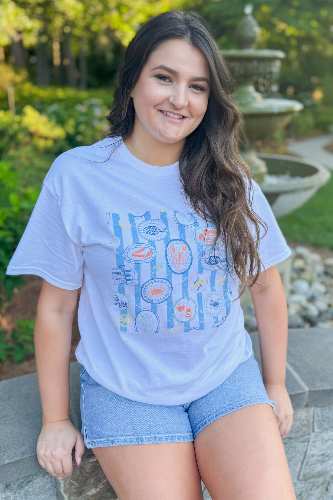 Front view of woman wearing Swagger Boutique’s White &amp; Blue Printed Graphic T-Shirt with jean shorts outside at a park.