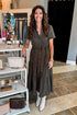 A woman stands in a boutique wearing a short-sleeve, button-front, tiered midi dress in dark olive brown, paired with white pointed-toe boots. The boutique interior features white display shelves with handbags and accessories on the left, and a black curtain in the background.