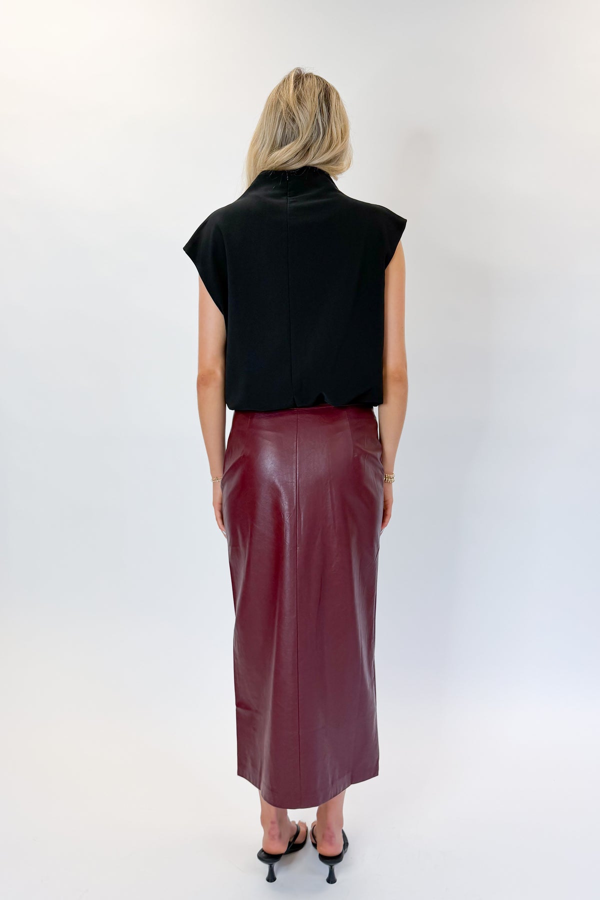 Back view of woman wearing Swagger Boutique’s Wine Faux Leather Slit Midi Skirt with black sweater against white studio background.