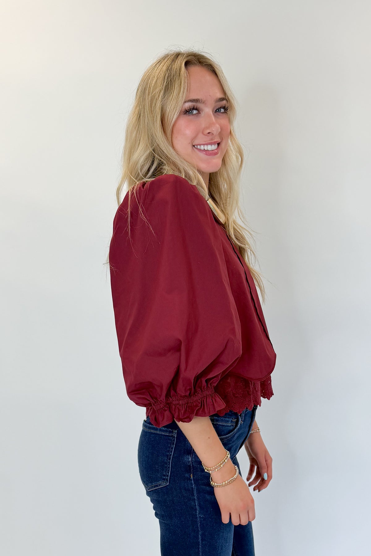 Side view of woman wearing Swagger Boutique’s Wine Lace Trim Top by Entro with jeans against white studio background.