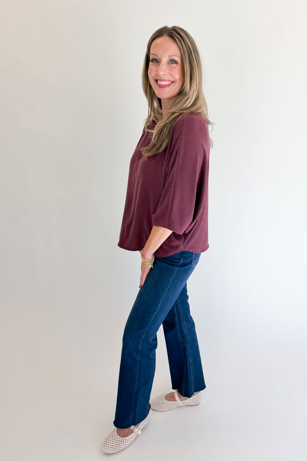 Side view of woman wearing Swagger Boutique’s Wine Ribbed Dolman Sleeve Top with jeans against white studio background.