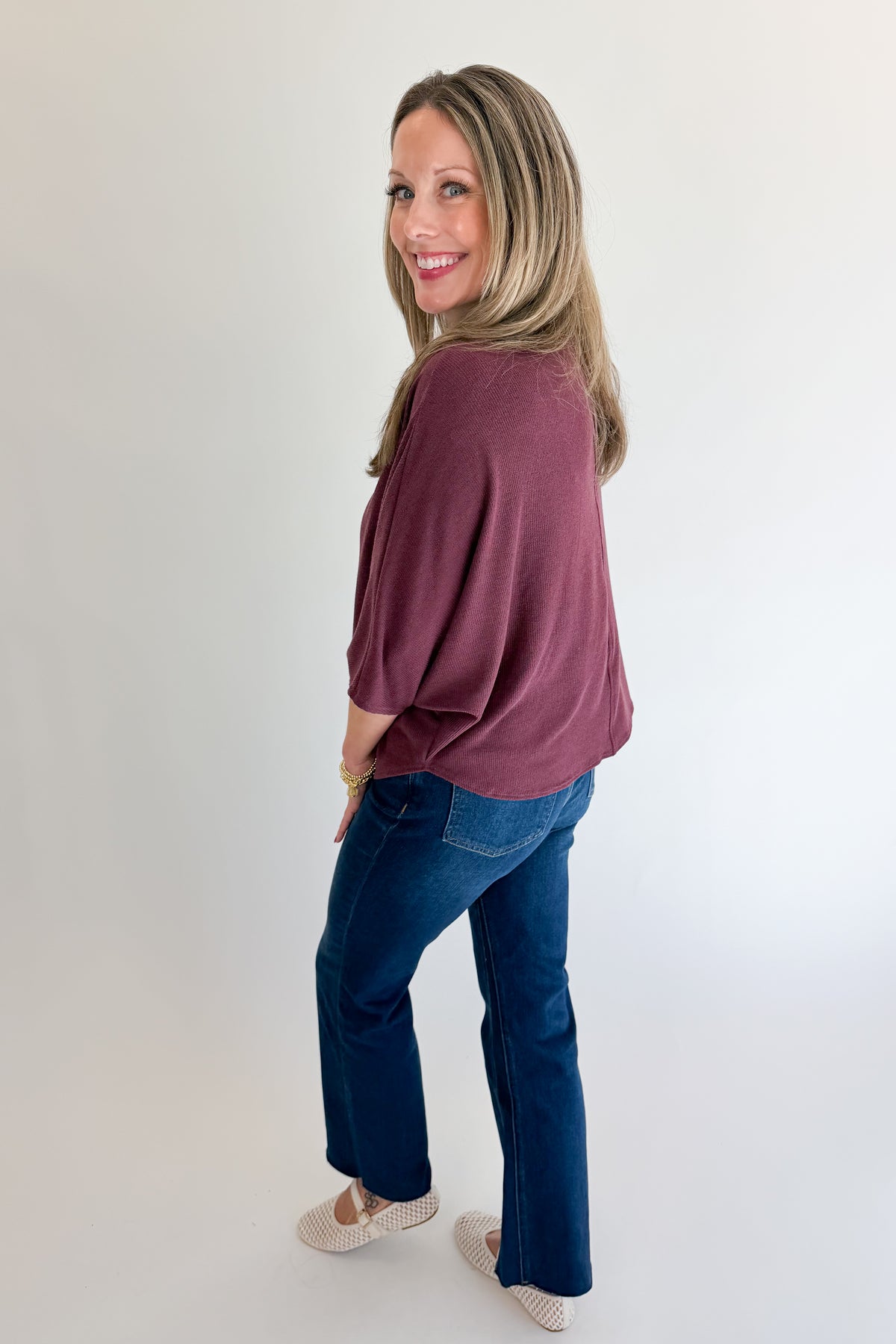 Side view of woman wearing Swagger Boutique’s Wine Ribbed Dolman Sleeve Top with jeans against white studio background.