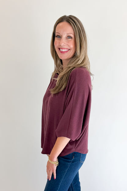 Side view of woman wearing Swagger Boutique’s Wine Ribbed Dolman Sleeve Top with jeans against white studio background.