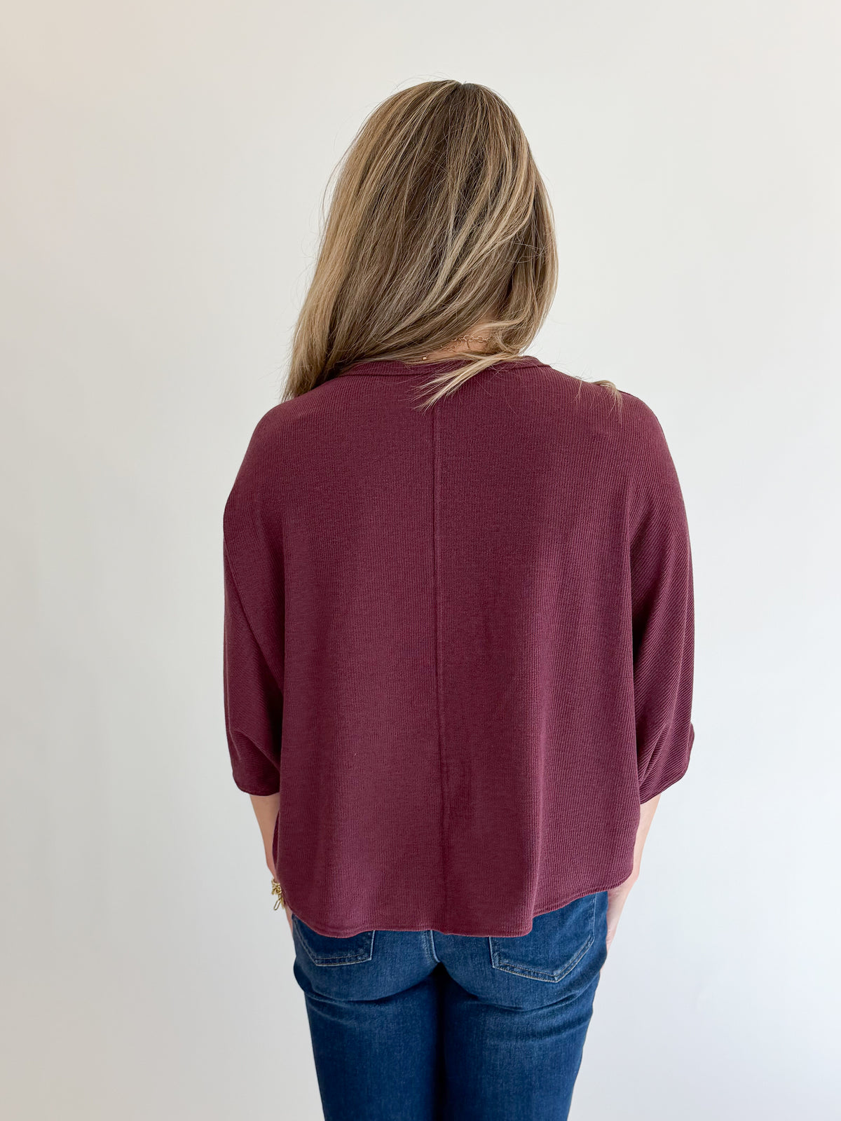 Back view of woman wearing Swagger Boutique’s Wine Ribbed Dolman Sleeve Top with jeans against white studio background.
