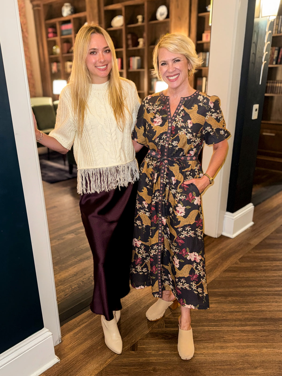 Front view of women wearing Swagger Boutique’s Wine Satin Maxi Skirt with Cream Fringe Short Sleeve Sweater and Navy Animal Print Midi Dress standing outside a den.